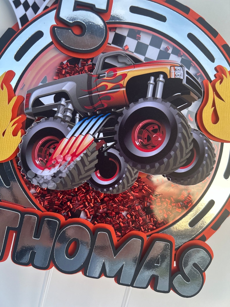 May include: A silver and red monster truck cake topper with the number 5 and the name Thomas. The truck has flames on the side and is surrounded by red confetti.