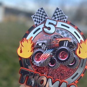 May include: A red, white, and black monster truck cake topper with a silver number 5, checkered flags, and the name "Thomas" in silver letters.