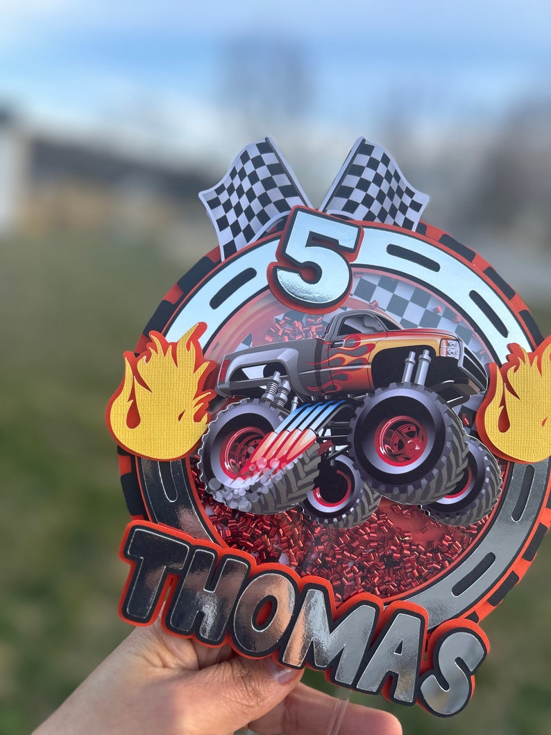 May include: A red, white, and black monster truck cake topper with a silver number 5, checkered flags, and the name "Thomas" in silver letters.