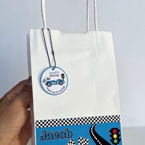 Puede incluir: Bolsa de regalo de papel blanco con un diseño de coche de carreras azul. La bolsa presenta un borde de bandera a cuadros y el nombre "Jacob". Una etiqueta circular dice "¡Cumpleaños de Jacob!" y "¡Gracias por venir!"