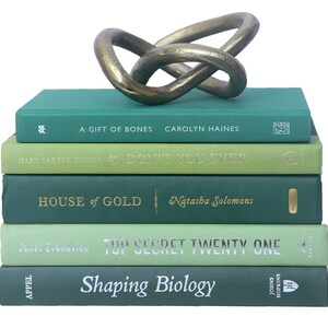 May include: A stack of five green hardcover books with gold lettering on the spines. The books are titled "A Gift of Bones", "Don't You Ever", "House of Gold", "Top Secret Twenty One", and "Shaping Biology". A gold metal knot sculpture sits on top of the books.