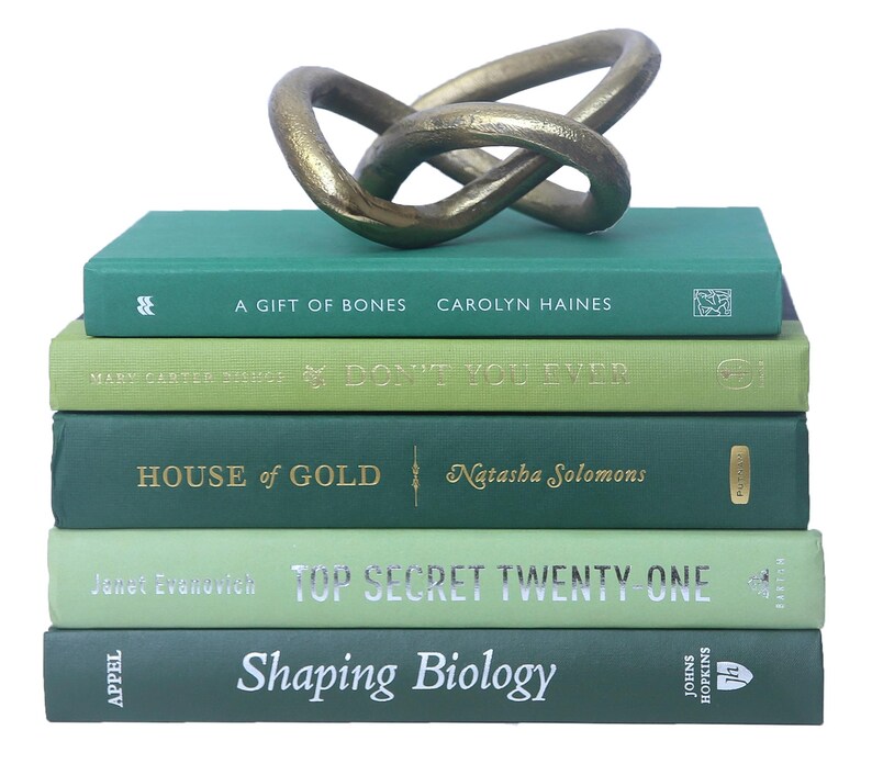 May include: A stack of five green hardcover books with gold lettering on the spines. The books are titled "A Gift of Bones", "Don't You Ever", "House of Gold", "Top Secret Twenty One", and "Shaping Biology". A gold metal knot sculpture sits on top of the books.
