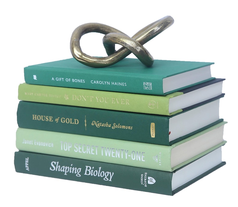 May include: A stack of five green hardcover books with gold lettering on the spines. The top book has the title "A Gift of Bones" and the author "Carolyn Haines". The second book has the title "Don't You Ever". The third book has the title "House of Gold" and the author "Natasha Solomons". The fourth book has the title "Top Secret Twenty-One". The fifth book has the title "Shaping Biology" and the publisher "Johns Hopkins". A gold metal sculpture of a knot is on top of the books.