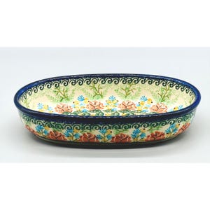 May include: An oval ceramic baking dish featuring a cream-colored base with a hand-painted floral pattern of orange roses, blue accents, and green foliage. The rim is decorated with a dark blue swirl border, and the exterior sides feature a repeating pattern of roses and blue floral motifs.