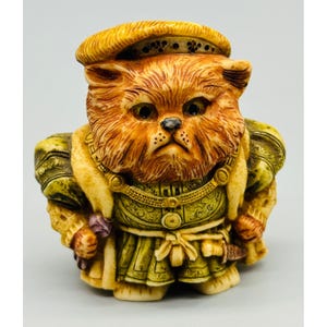 May include: A small figurine of a fluffy orange cat dressed in historical attire, including a golden beret, a green patterned tunic with a tied waist, and a decorative chain necklace. The cat has a serious expression and textured fur details.