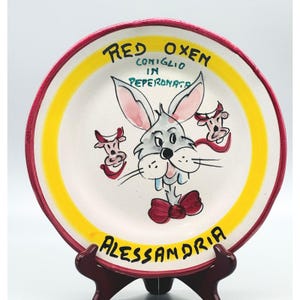 May include: A round ceramic plate with a red rim and a yellow inner border. The center features a hand-painted illustration of a rabbit with a red bow tie, flanked by two small ox heads. Text reads RED OXEN, CONIGLIO IN PEPERONATA, and ALESSANDRIA.