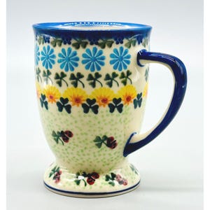May include: A ceramic mug with a flared base and a curved handle, featuring a cream-colored background decorated with patterns of blue daisies, green shamrocks, yellow flowers, and small red ladybugs, finished with a solid blue rim and handle.