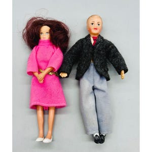 May include: Two dolls standing side by side. The doll on the left has dark reddish-brown hair and wears a bright pink long-sleeved dress with a gold belt. The doll on the right has short light hair and wears a dark gray blazer, red tie, and light gray pants.