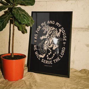 May include: A framed rectangular print featuring a cream-colored tiger illustration centered on a black background. The text "AS FOR ME AND MY HOUSE WE WILL SERVE THE LORD" circles the tiger, with "JOSHUA 24:15" printed in small text at the bottom.