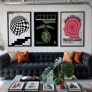 Puede incluir: Un salón con un sofá gris oscuro con botones y cojines naranjas y beige. Tres impresiones artísticas enmarcadas cuelgan sobre el sofá, con diseños geométricos y botánicos. Una mesa de centro con tapa de cristal exhibe fotos enmarcadas y una planta en maceta.