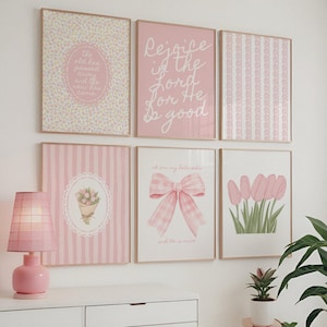 May include: A collection of six framed art prints in shades of pink and white, with floral, striped, and text designs. A pink lamp sits on a white dresser below the prints, with a potted plant to the right.
