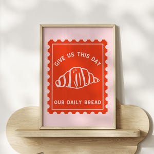 May include: A framed art print featuring a red postage stamp design with a white croissant illustration and the text "GIVE US THIS DAY OUR DAILY BREAD" on a pink background. The print is displayed on a cloud-shaped wooden shelf.