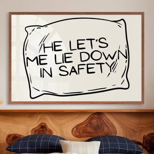 Può includere: Una stampa incorniciata con un disegno di cuscino e la scritta "HE LET'S ME LIE DOWN IN SAFETY" in nero. L'opera d'arte è su uno sfondo color crema, incorniciata in una cornice di legno marrone. La stampa è sopra un letto con cuscini e biancheria da letto.