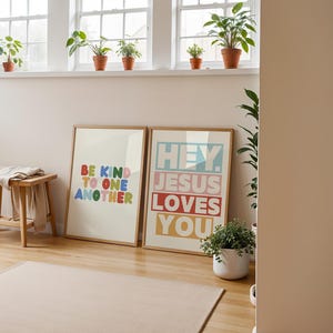 May include: Two framed art prints with inspirational messages. One reads "BE KIND TO ONE ANOTHER" in colorful letters. The other says "HEY. JESUS LOVES YOU" in a similar style. The prints are displayed on a wooden floor.
