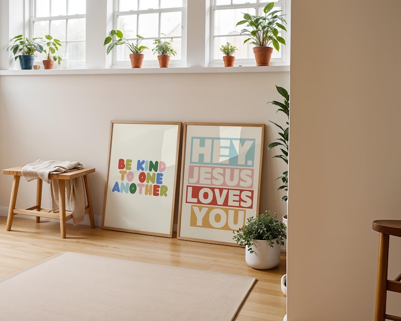 May include: Two framed art prints with inspirational messages. One reads "BE KIND TO ONE ANOTHER" in colorful letters. The other says "HEY. JESUS LOVES YOU" in a similar style. The prints are displayed on a wooden floor.
