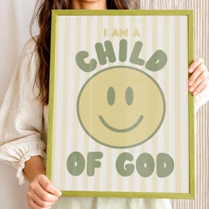 May include: A framed print with a green border features the text "I AM A CHILD OF GOD" in green lettering. A large yellow smiley face is centered below the word "CHILD". The background has vertical stripes in light yellow and white.