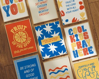 May include: A collection of framed art prints with inspirational quotes and phrases. The prints feature bold typography and colorful designs, including phrases like "LOVES YOU" and "COOL KIDS PRAY HERE". The artwork is displayed on a wooden surface.