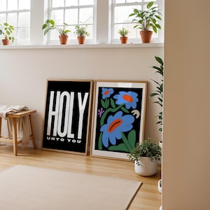 May include: Two framed prints on a white wall. The print on the left is black with white text that reads "HOLY UNTO YOU". The print on the right features a colourful illustration of blue flowers with orange centres on a black background.