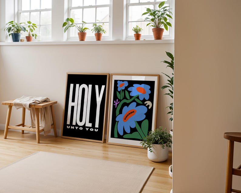 May include: Two framed prints on a white wall. The print on the left is black with white text that reads "HOLY UNTO YOU". The print on the right features a colourful illustration of blue flowers with orange centres on a black background.