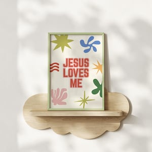 May include: A framed print with the text "JESUS LOVES ME" in red, set against a cream background with colorful abstract shapes. The frame is light green, and the print rests on a cloud-shaped wooden shelf.