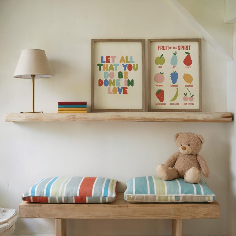 May include: A cozy interior scene featuring a wooden shelf with framed art prints. One print reads "LET ALL THAT YOU DO BE DONE IN LOVE" in colorful letters. The other print displays "FRUIT OF THE SPIRIT". Below, striped pillows and a teddy bear rest on a wooden bench.