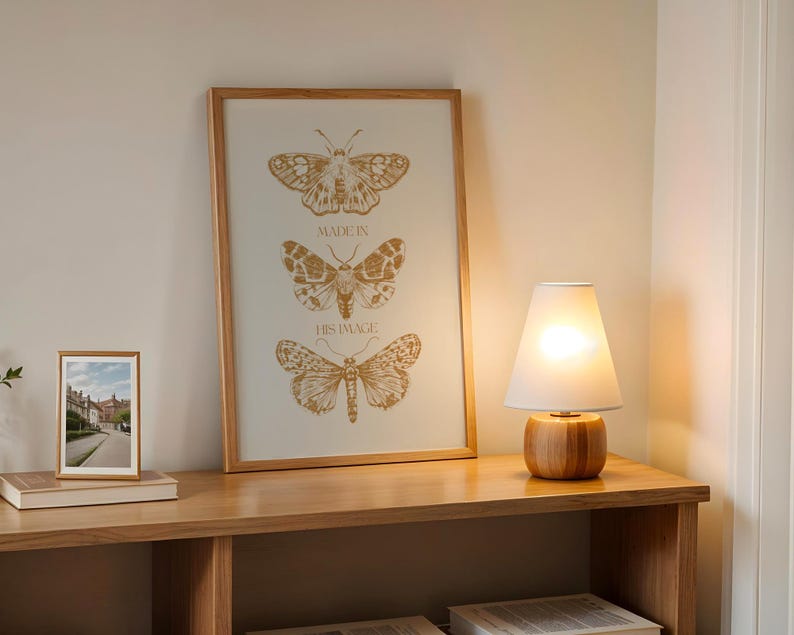 May include: A wooden framed print featuring three detailed butterfly illustrations in shades of gold, with the text "MADE IN HIS IMAGE". A small framed photo and a wooden lamp with a white shade sit on a wooden shelf.