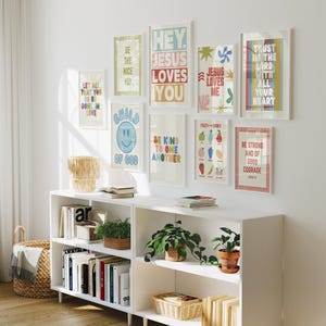 May include: A white wall displays framed art prints with inspirational quotes and colorful designs. A white bookshelf below holds books, plants, and decorative baskets. The prints include phrases like "Hey Jesus Loves You" and "Be Strong and of Good Courage."