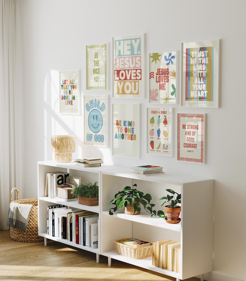 May include: A white wall displays framed art prints with inspirational quotes and colorful designs. A white bookshelf below holds books, plants, and decorative baskets. The prints include phrases like "Hey Jesus Loves You" and "Be Strong and of Good Courage."