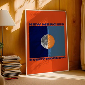 May include: A poster with an orange background and a blue and black rectangle in the centre. The rectangle has a half moon in it. The text "NEW MERCIES EVERY MORNING" is printed in blue on the poster.