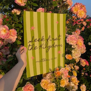 May include: A rectangular sign with vertical green stripes and the text "Seek first the Kingdom" and "matthew 6:33" in a handwritten style. Pink star accents are scattered across the sign. The sign is held in front of a backdrop of pink and yellow roses.