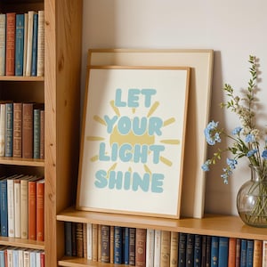 May include: A framed print with the words "LET YOUR LIGHT SHINE" in blue and yellow. The print is displayed on a wooden shelf with a bookshelf and a vase of blue flowers. The print has a light beige border.