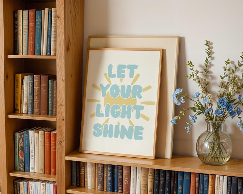 May include: A framed print with the words "LET YOUR LIGHT SHINE" in blue and yellow. The print is displayed on a wooden shelf with a bookshelf and a vase of blue flowers. The print has a light beige border.