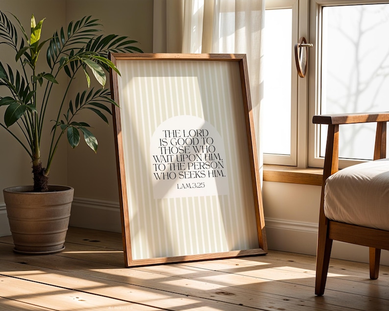 May include: A framed print with a wooden frame, featuring the text "THE LORD IS GOOD TO THOSE WHO WAIT UPON HIM, TO THE PERSON WHO SEEKS HIM. LAM325" in a serif font. The background has vertical stripes. A potted plant and a chair are in the background.