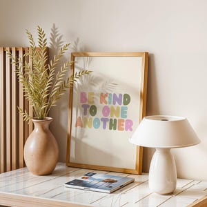May include: A framed art print with the words "BE KIND TO ONE ANOTHER" in colorful bubble letters. A white lamp, a vase with greenery, and a magazine are on a white and wood-toned table. The art print is in a light wood frame.