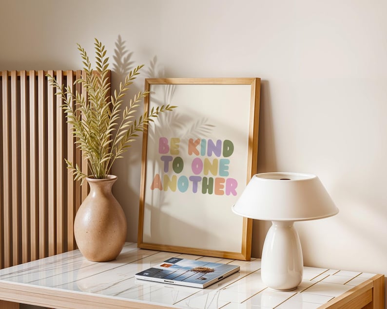 May include: A framed art print with the words "BE KIND TO ONE ANOTHER" in colorful bubble letters. A white lamp, a vase with greenery, and a magazine are on a white and wood-toned table. The art print is in a light wood frame.