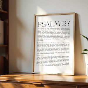May include: A framed print with the text of Psalm 27 in black font. The print is in a light wood frame and is displayed on a wooden surface. A small potted plant is visible on the right side of the image.