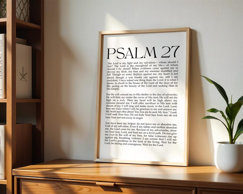 May include: A framed print with the text of Psalm 27 in black font. The print is in a light wood frame and is displayed on a wooden surface. A small potted plant is visible on the right side of the image.