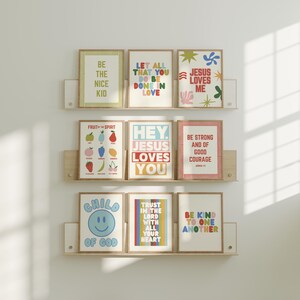 May include: Nine framed art prints with inspirational quotes and colorful designs are displayed on wooden shelves. The prints feature phrases like "Be the Nice Kid," "Jesus Loves Me," and "Trust in the Lord." The artwork includes a variety of fonts and illustrations.