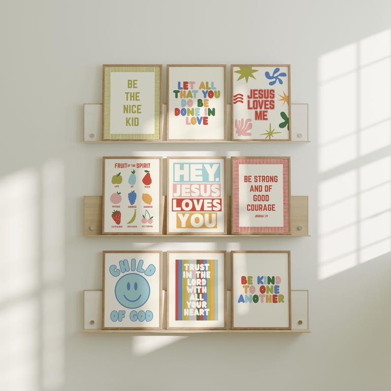 May include: Nine framed art prints with inspirational quotes and colorful designs are displayed on wooden shelves. The prints feature phrases like "Be the Nice Kid," "Jesus Loves Me," and "Trust in the Lord." The artwork includes a variety of fonts and illustrations.