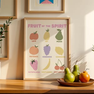 May include: A framed print titled "Fruit of the Spirit" with colorful illustrations of fruits representing virtues. The print is in a light wood frame and is displayed on a wooden surface. Two green pears and an orange fruit sit on a wooden plate.