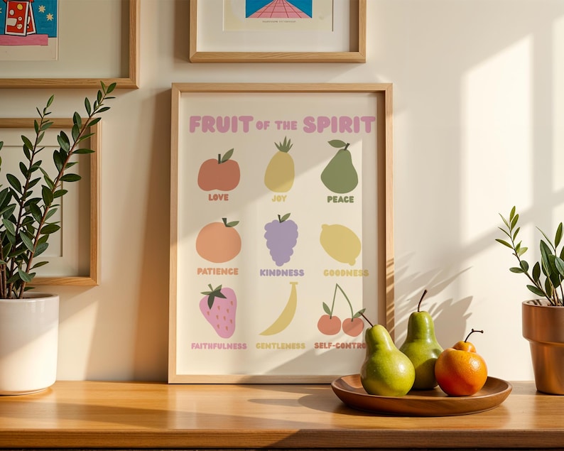May include: A framed print titled "Fruit of the Spirit" with colorful illustrations of fruits representing virtues. The print is in a light wood frame and is displayed on a wooden surface. Two green pears and an orange fruit sit on a wooden plate.
