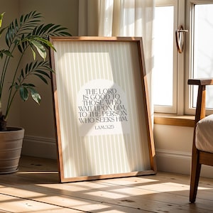 May include: A framed print with a wooden frame, featuring the text "THE LORD IS GOOD TO THOSE WHO WAIT UPON HIM, TO THE PERSON WHO SEEKS HIM. LAM325" in a serif font. The background has vertical stripes. A potted plant and a chair are in the background.