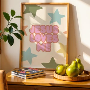 May include: A framed art print with the text "JESUS LOVES ME" in pink bubble letters, surrounded by pastel stars. The artwork is displayed on a wooden surface with a wooden tray holding green pears and a floral-covered notebook.