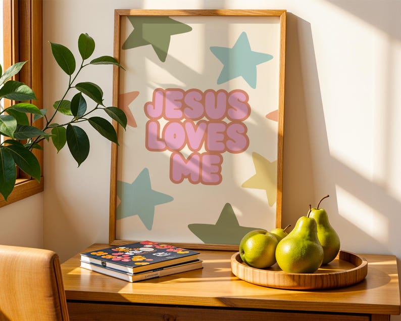 May include: A framed art print with the text "JESUS LOVES ME" in pink bubble letters, surrounded by pastel stars. The artwork is displayed on a wooden surface with a wooden tray holding green pears and a floral-covered notebook.