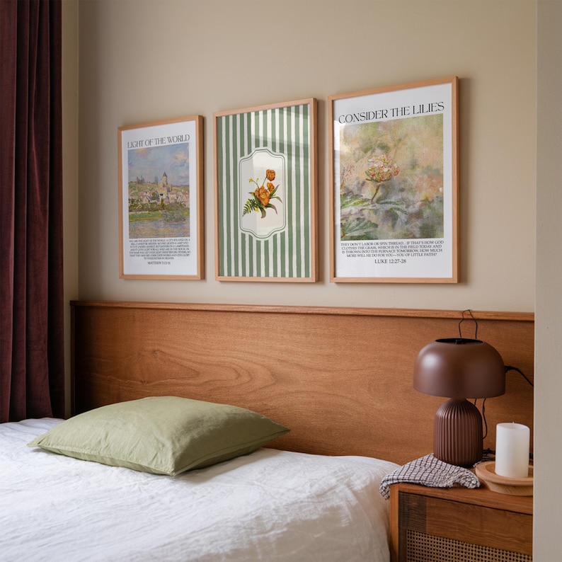 May include: Three framed art prints hang above a bed. The prints feature botanical and landscape imagery with text. The middle print has a green and white striped background with a floral design. A green pillow rests on the white bedsheets. A brown lamp and candle sit on the nightstand.