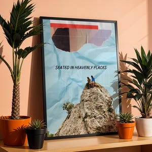 May include: A black framed poster with a blue background and a ripped paper effect. The poster features a photo of two people sitting on a rocky mountaintop. The text "Seated in Heavenly Places" is printed on the poster.