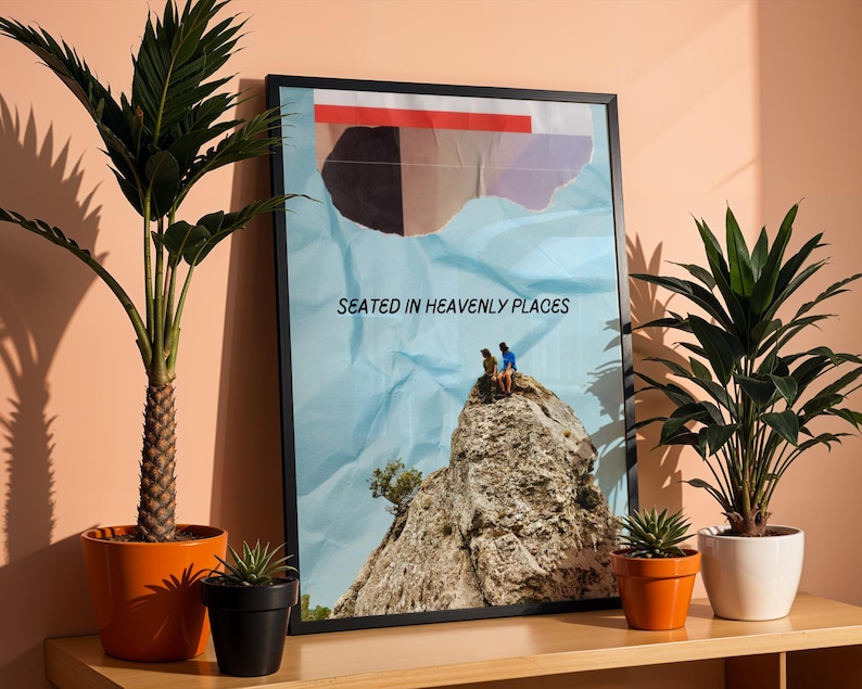 May include: A black framed poster with a blue background and a ripped paper effect. The poster features a photo of two people sitting on a rocky mountaintop. The text "Seated in Heavenly Places" is printed on the poster.