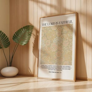 May include: A framed print with the text "THE LORD IS FAITHFUL" and a floral pattern in beige and pink. The print is displayed on a light wood surface, next to a potted plant with large green leaves.