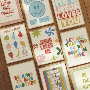 May include: A collection of framed art prints with inspirational quotes and colorful designs. The prints feature phrases like "Jesus Loves Me," "Be Kind to One Another," and "Fruit of the Spirit." The artwork is displayed on a wooden surface.