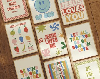May include: A collection of framed art prints with inspirational quotes and colorful designs. The prints feature phrases like "Jesus Loves Me," "Be Kind to One Another," and "Fruit of the Spirit." The artwork is displayed on a wooden surface.